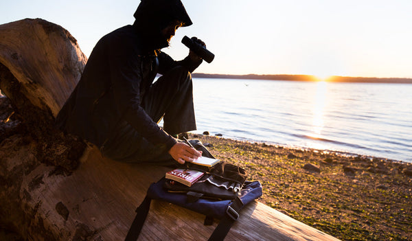 Person sitting on a log at the beach, holding a pair of binoculars and taking notes. The Large Cafe Bag with its Freudian Slip sits next to them.